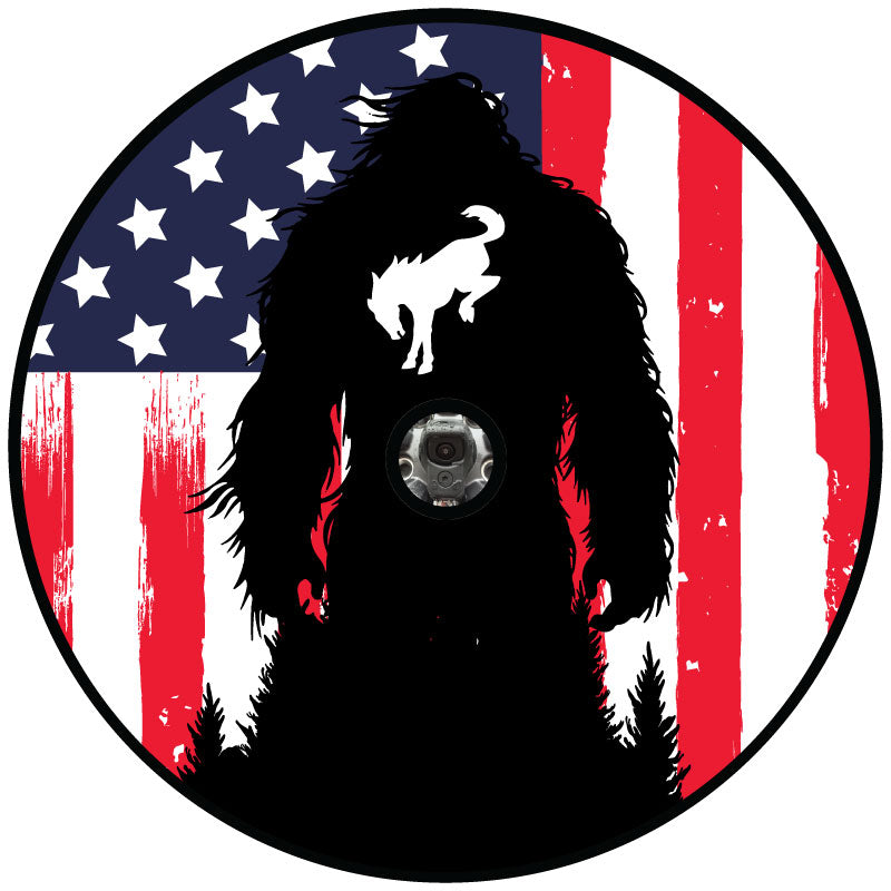 A spare tire cover design mockup of a rustic vertical  American flag background in red white and blue with a silhouette of Sasquatch and the bucking Bronco icon on bigfoot's chest with a hole to show placement if you have a backup camera on your spare tire cover model.