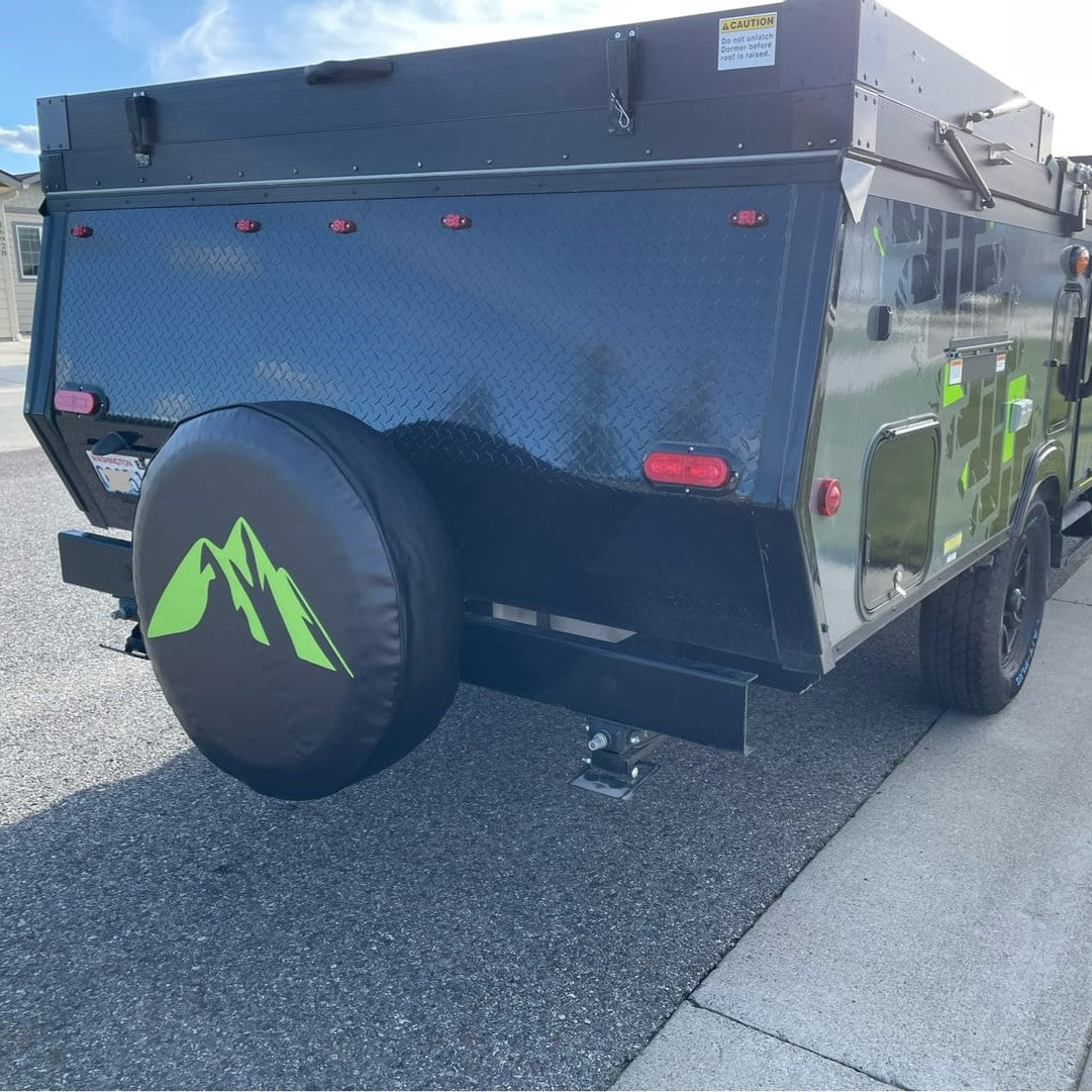 Black pop up camper from the back parked on the street showing a black vinyl spare tire cover design of a simple lime green mountain silhouette graphic.