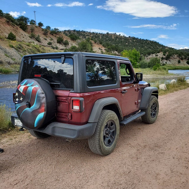 Backside view of a maroon red Jeep with a patriotic waving American flag spare tire cover as it's parked next to a river in the hills.