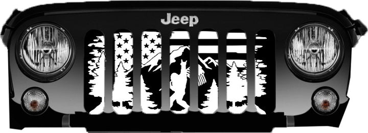 Close up view of front end of a Jeep Wrangler grille showing a Jeep grille insert design of an American flag background, mountains and a forest, with Bigfoot or Sasquatch walking by showing a peace sign holding the American flag.