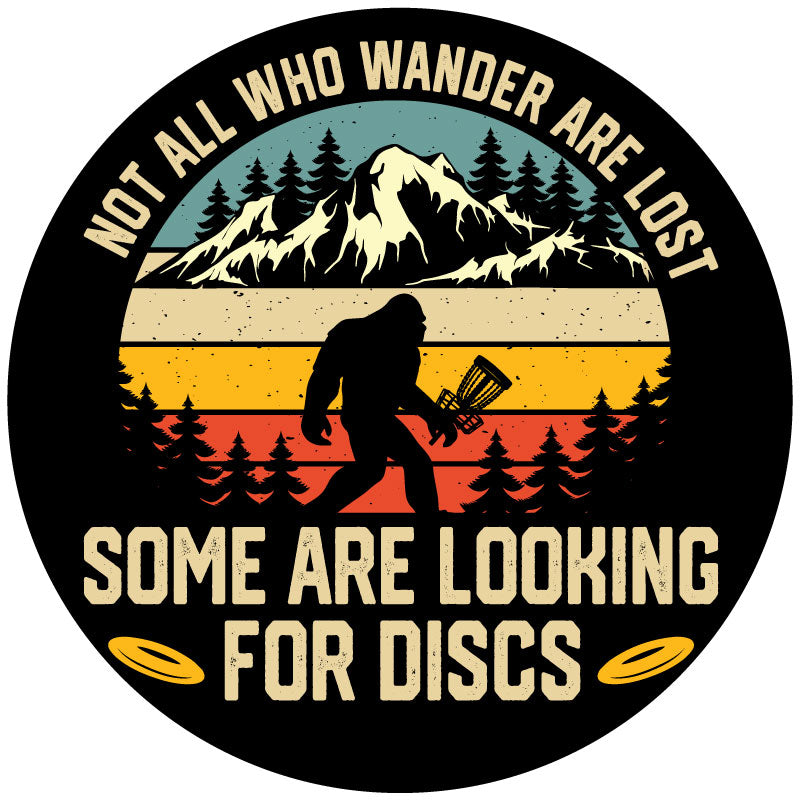A funny spare tire cover design of sasquatch walking through the wilderness holding a disc golf basket and the saying not all who wonder are lost some are looking for discs on black background