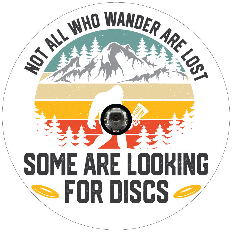 A funny spare tire cover design of sasquatch walking through the wilderness holding a disc golf basket and the saying not all who wonder are lost some are looking for discs on white background with JL back up camera