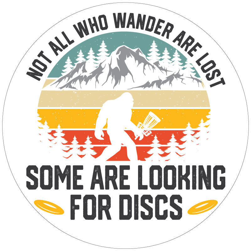 A funny spare tire cover design of sasquatch walking through the wilderness holding a disc golf basket and the saying not all who wonder are lost some are looking for discs on white background